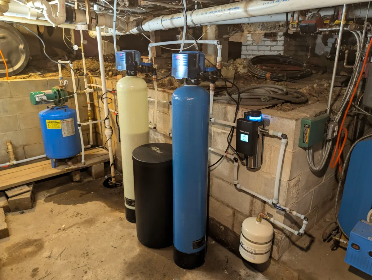 Whole-home water softener system with treatment tanks, control valves, filtration equipment, and connected plumbing in a basement utility area.